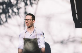 Ein Mann mit Brille und Schürze steht vor einer weißen Wand im Sonnenlicht.